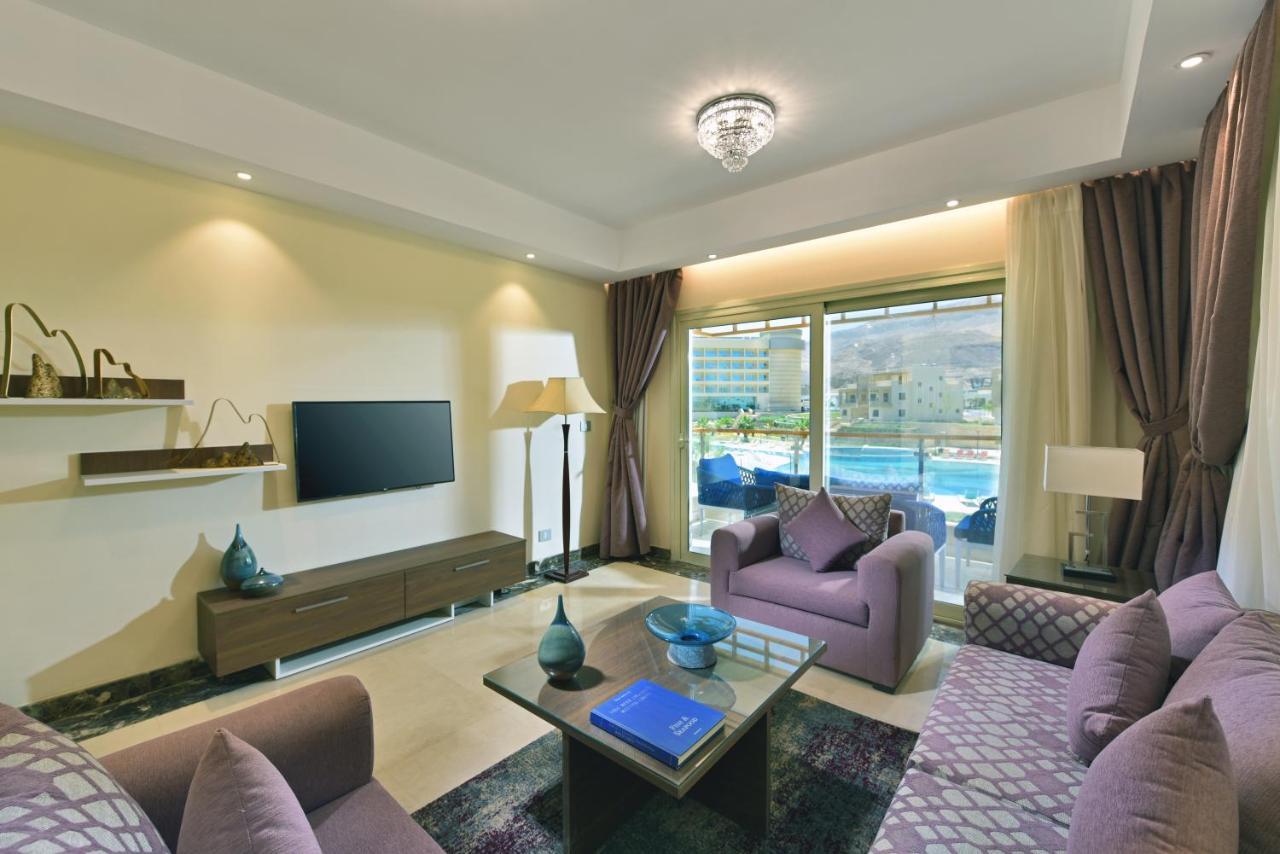 Tolip Resort El Galala Majestic Ain Suchna Exterior foto The photo shows a modern living room that features a stylish and cozy design. There are two purple sofas with decorative cushions arranged around a coffee table. The coffee table has a blue book and a decorative item on it. A flat-screen TV is mounte