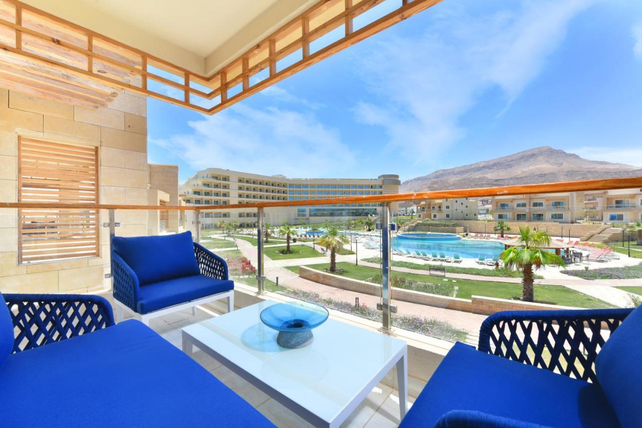 Tolip Resort El Galala Majestic Ain Suchna Exterior foto The photo showcases a balcony view featuring two blue chairs and a table with a decorative bowl. The balcony overlooks a landscape that includes a swimming pool and manicured lawns. In the background, there are multiple buildings and palm trees, all 