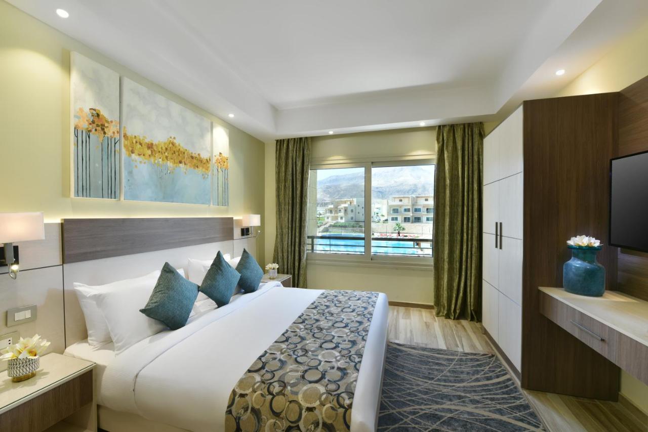 Tolip Resort El Galala Majestic Ain Suchna Exterior foto The photo shows a modern hotel room. It features a large bed with white bedding and several decorative pillows in shades of teal. Above the bed, there are two pieces of wall art with a landscape theme. The room has a large window with green curtains,