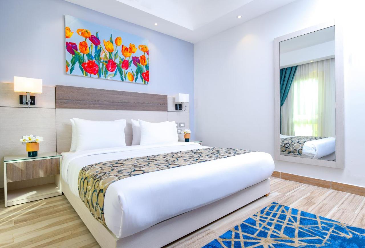 Tolip Resort El Galala Majestic Ain Suchna Exterior foto The photo shows a modern hotel bedroom. It features a large bed with white linens and a decorative throw blanket. The room has light wooden flooring and is bright, with ample natural light coming from a window. There is a colorful painting of flowers