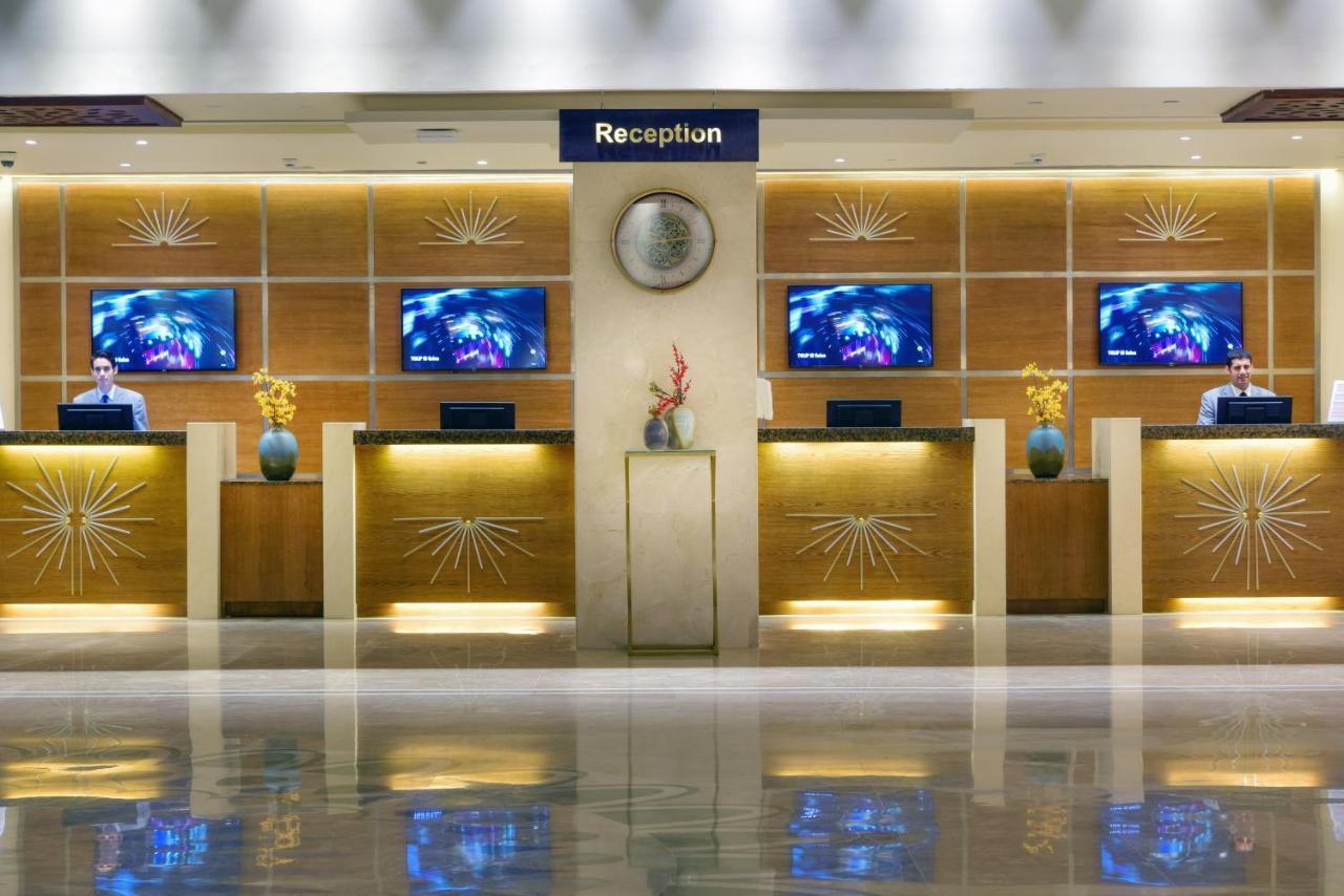 Tolip Resort El Galala Majestic Ain Suchna Exterior foto The image shows a hotel reception area. There are several reception desks lined up against a wall with decorative paneling. Each desk has a staff member, and above the desks, there are screens displaying information. A clock is visible on the wall, a