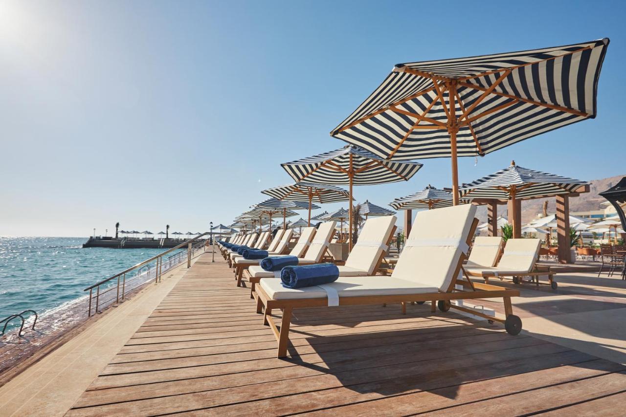 Tolip Resort El Galala Majestic Ain Suchna Exterior foto The photo depicts a serene beachside setting featuring a row of sun loungers arranged on a wooden deck. Each lounger is equipped with a folded towel and shaded by striped umbrellas. In the background, the water sparkles under bright sunlight, and the