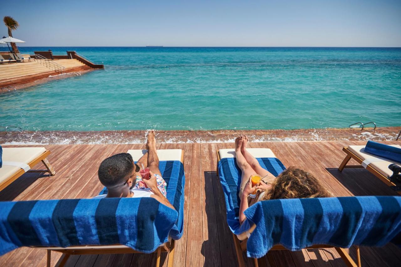 Tolip Resort El Galala Majestic Ain Suchna Exterior foto The photo depicts two people relaxing on lounge chairs by the waterfront. They are positioned on a wooden deck with a view of the clear, turquoise water. Both individuals are reclining, enjoying the sun, and each has a drink in hand. The scene convey