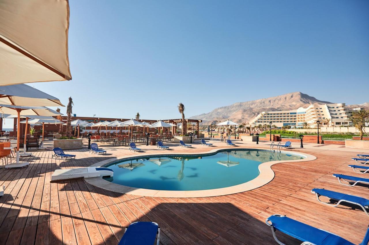 Tolip Resort El Galala Majestic Ain Suchna Exterior foto The photo shows a swimming pool area surrounded by sun loungers and umbrellas. The pool has a unique, curvy shape and is situated on a wooden deck. In the background, there are several buildings, likely part of a resort or hotel, and a mountainous la