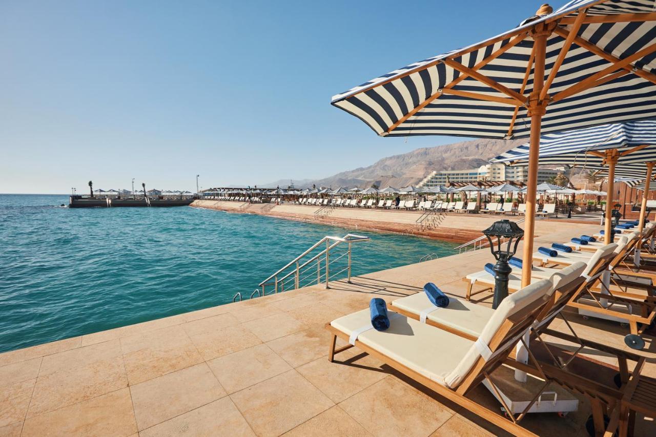 Tolip Resort El Galala Majestic Ain Suchna Exterior foto The photo shows a scenic beachside area with clear blue water. In the foreground, there are several sun loungers set up on a tiled surface, with blue towels neatly rolled on each one. The loungers are shaded by striped umbrellas. In the distance, a w
