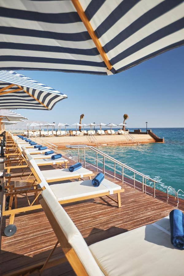 Tolip Resort El Galala Majestic Ain Suchna Exterior foto The photo depicts a serene outdoor area by the waterfront. There are several lounge chairs arranged in a row, each equipped with blue towels. Large, striped umbrellas provide shade over some of the chairs. In the background, there is a wooden deck th