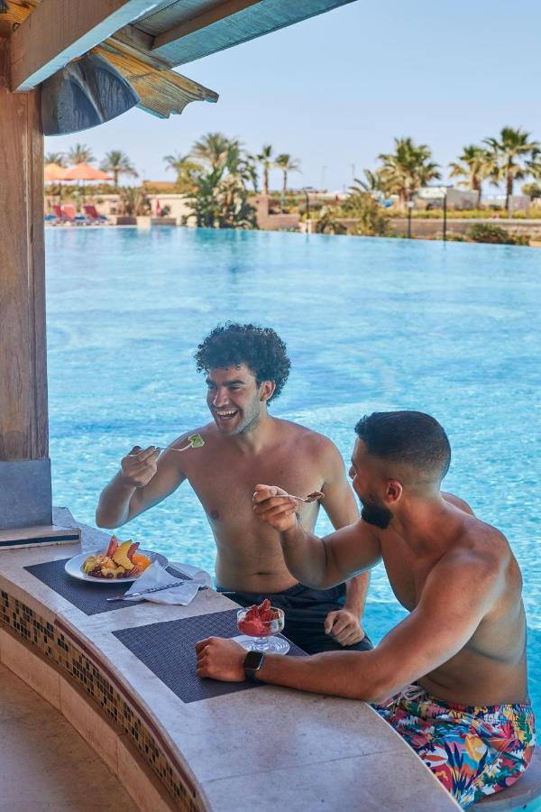 Tolip Resort El Galala Majestic Ain Suchna Exterior foto The photo features two young men enjoying a meal together. They are sitting at a poolside table with a clear blue swimming pool in the background. The men are shirtless, suggesting a casual, relaxed atmosphere, likely in a warm environment. One man i