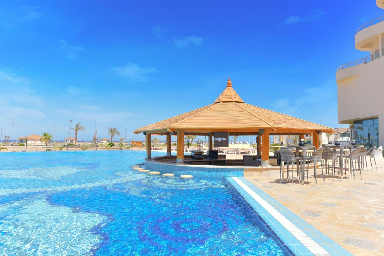 Tolip Resort El Galala Majestic Ain Suchna Exterior foto The photo shows a beautiful outdoor setting featuring a large swimming pool with clear blue water. In the center, there is a thatched-roof gazebo, which serves as a shaded seating area. Surrounding the gazebo are lounge chairs and a bar area with hig