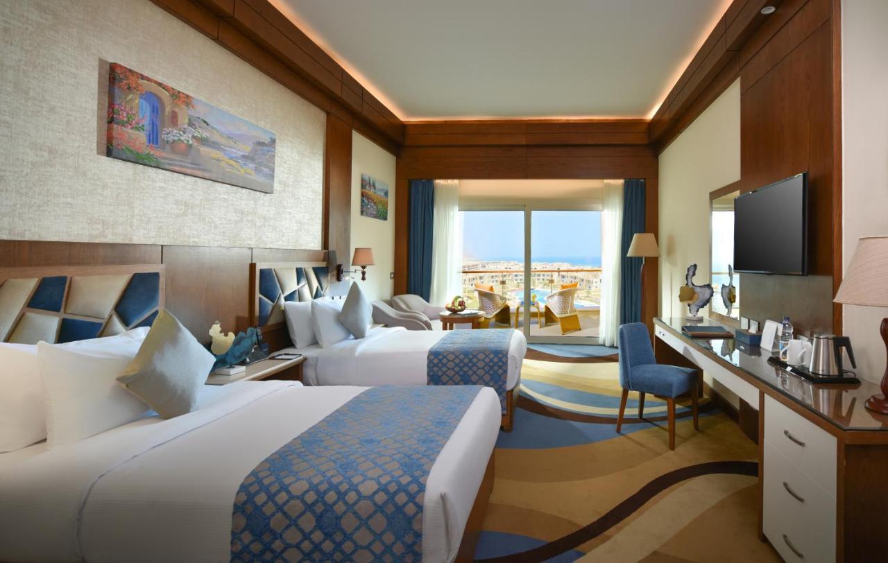 Tolip Resort El Galala Majestic Ain Suchna Exterior foto The photo shows a well-decorated hotel room featuring two beds with decorative pillows. The room has wooden accents and modern furnishings, including a desk, a television, and lamps. Large windows flood the space with natural light, offering a view o