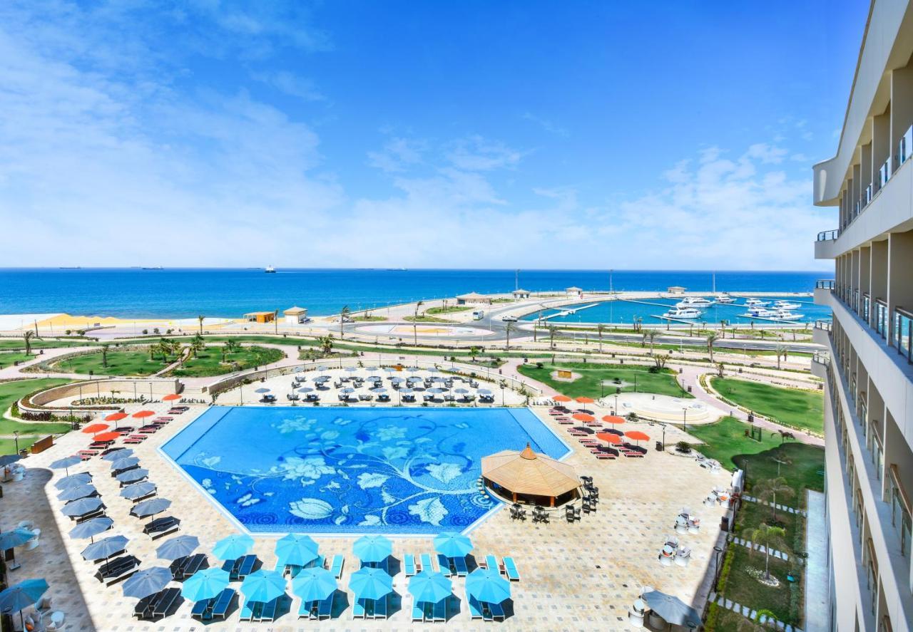 Tolip Resort El Galala Majestic Ain Suchna Exterior foto The photo shows a beautiful outdoor view of a resort area featuring a large swimming pool with a decorative underwater design. Surrounding the pool are numerous sun loungers and umbrellas in a variety of colors. In the background, there is a scenic c