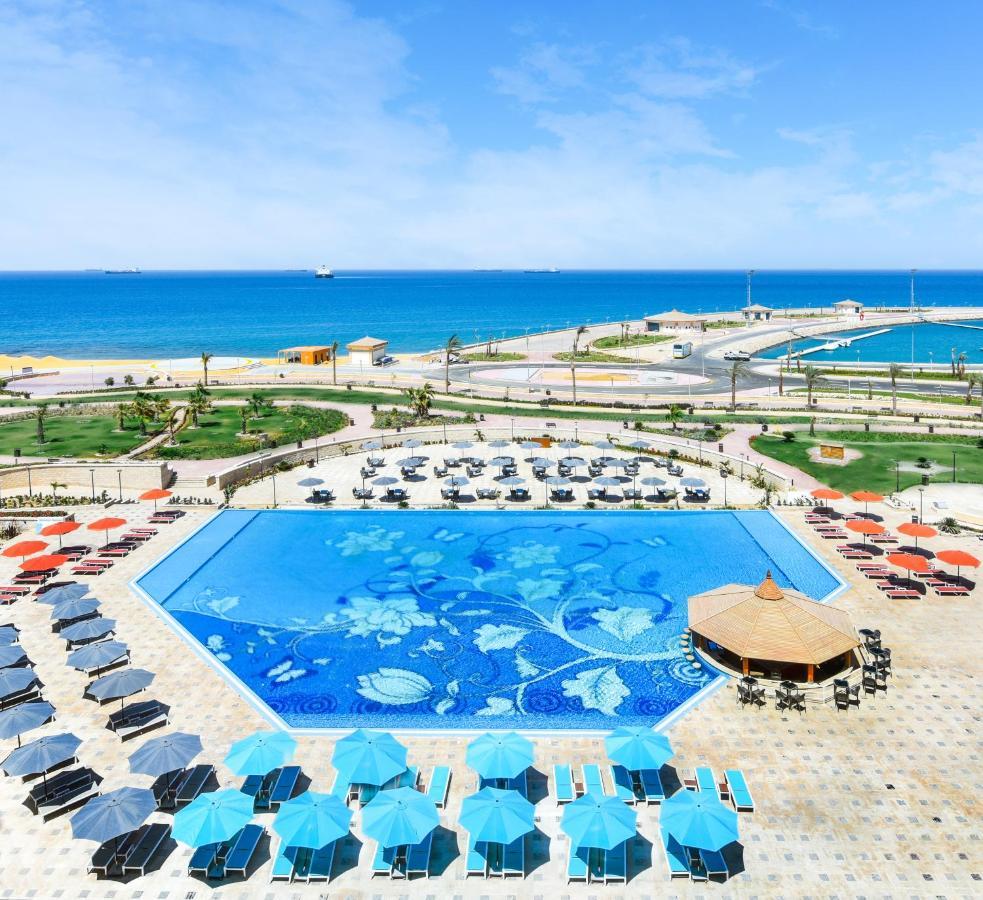 Tolip Resort El Galala Majestic Ain Suchna Exterior foto The photo features a large swimming pool with a decorative design on its surface, surrounded by sun loungers and umbrellas. In the background, there is a view of the sea, with a clear blue sky and a few scattered clouds. The area is well-maintained, 