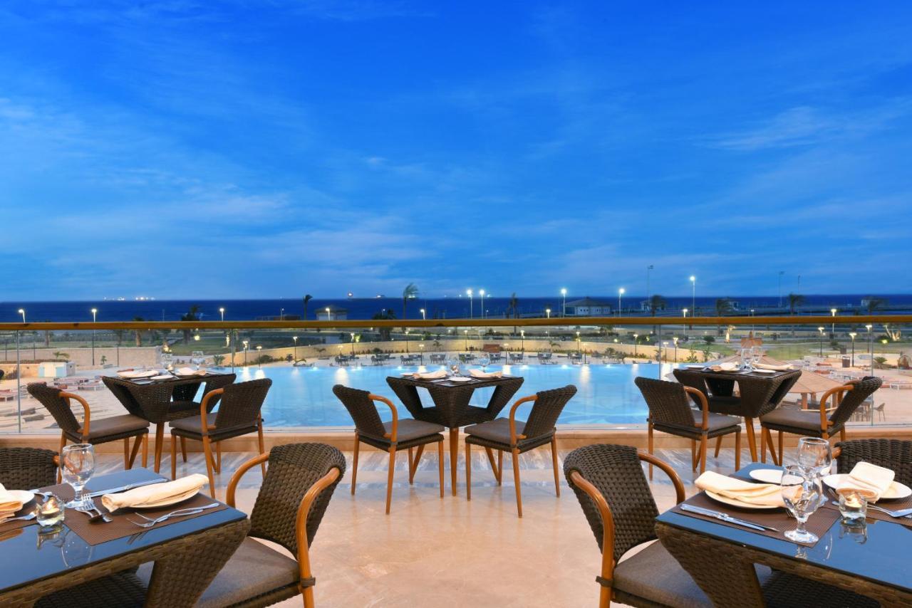 Tolip Resort El Galala Majestic Ain Suchna Exterior foto The photo shows an outdoor dining area with several neatly arranged tables and chairs. The tables are elegantly set with cutlery and plates, suggesting a fine dining experience. In the background, there is a large swimming pool that reflects the blue
