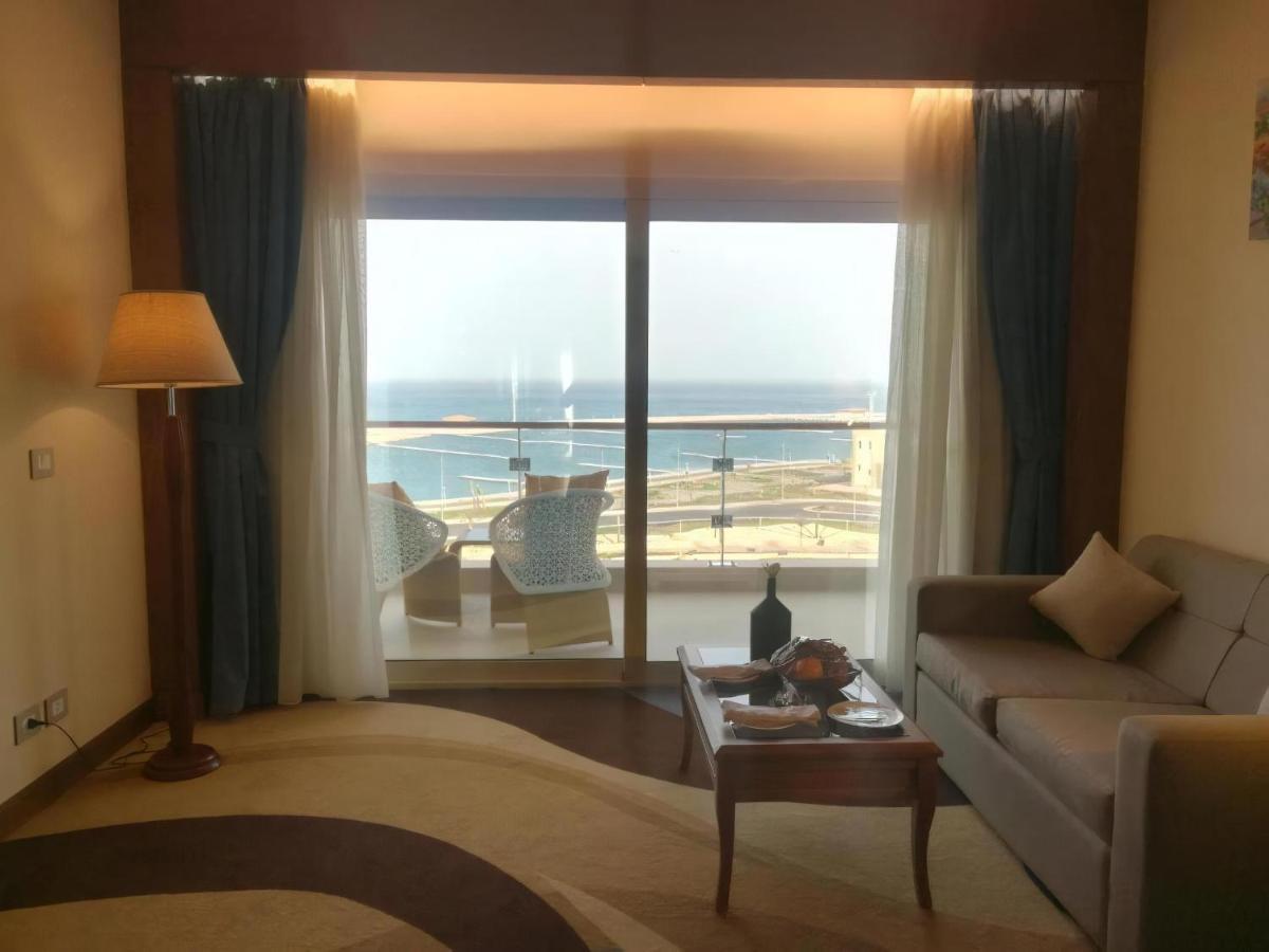 Tolip Resort El Galala Majestic Ain Suchna Exterior foto The photo shows a stylish interior of a room with a large window overlooking the sea. The window features sheer and thick curtains and is framed by a balcony with two chairs visible outside. Inside, there is a comfortable-looking sofa, a coffee table