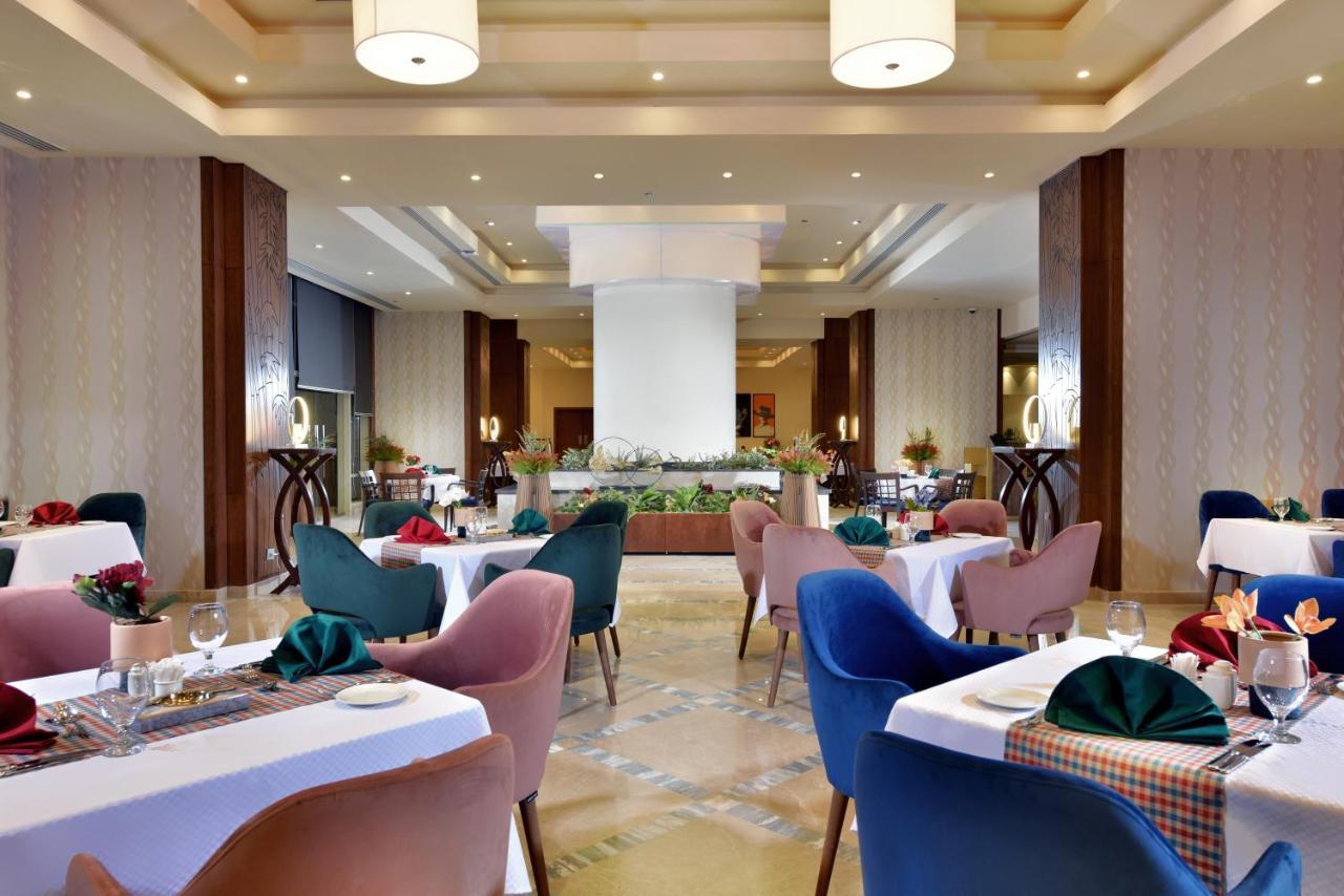 Tolip Resort El Galala Majestic Ain Suchna Exterior foto The photo shows a spacious and elegantly designed restaurant interior. The layout includes several round tables dressed in white tablecloths, each set with decorative plates, cutlery, and colorful napkins. The chairs are upholstered in a variety of c