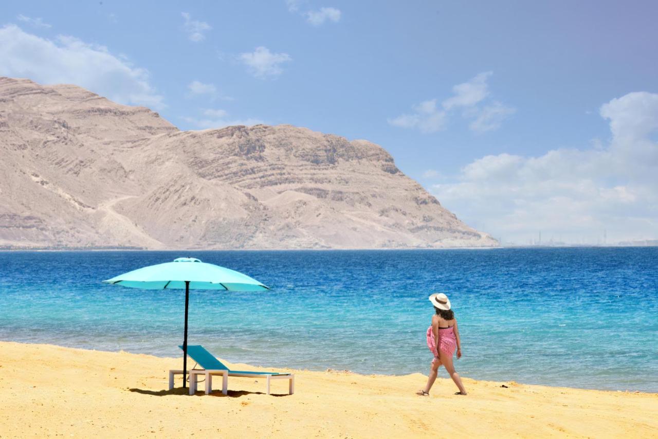 Tolip Resort El Galala Majestic Ain Suchna Exterior foto The photo depicts a serene beach scene. In the foreground, there is a sandy shore with a single beach chair and an umbrella in a light blue color. A person dressed in a pink outfit is walking along the shoreline, enjoying the beach atmosphere. In the